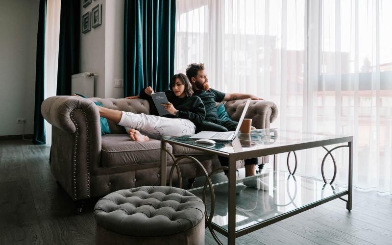 man sits on a couch and looks out the window while his wife lies against him reading a book