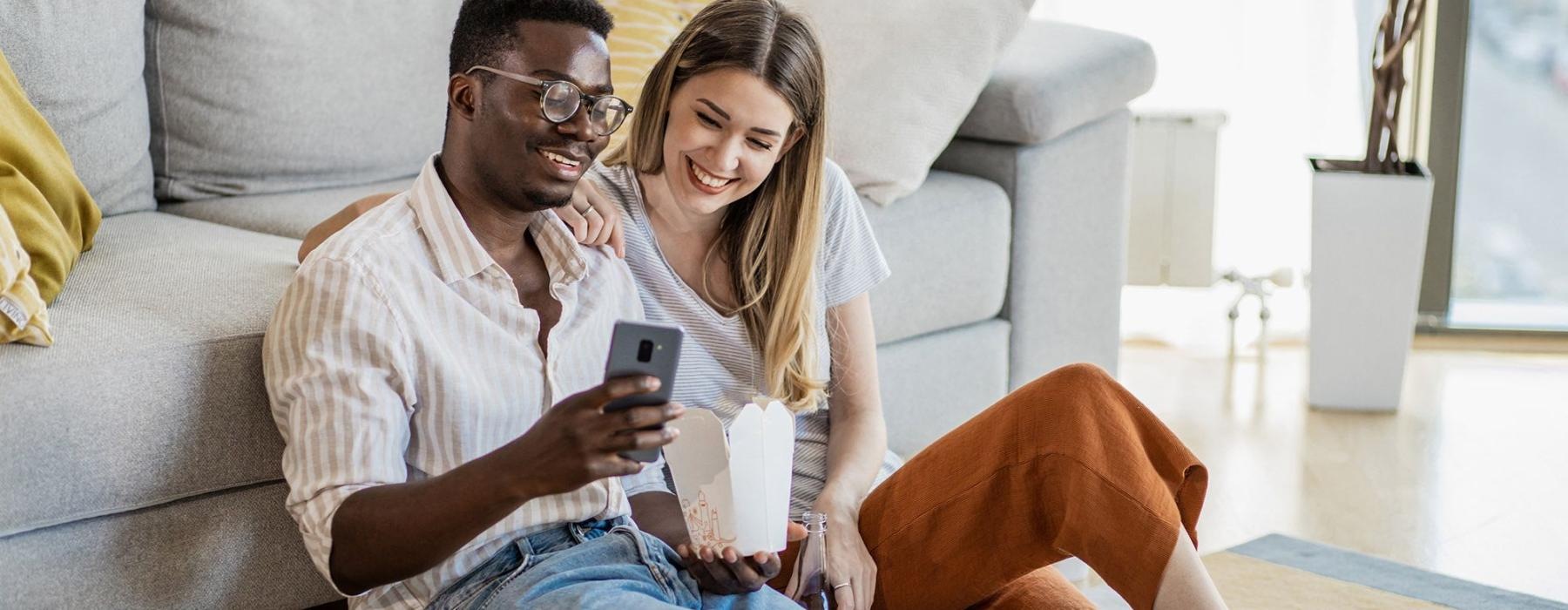 a man and woman with take out, sit against a couch on their living room floor and watch at their cell phone