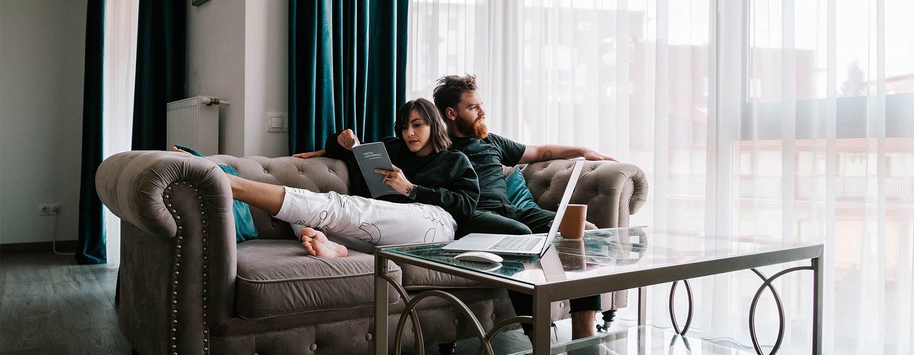 man sits on a couch and looks out the window while his wife lies against him reading a book